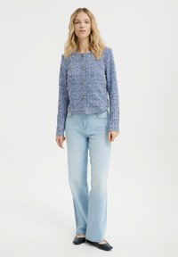 Giacca in tweed blu con colletto rotondo, chiusura frontale con bottoni e maniche lunghe, abbinata a jeans a zampa d'elefante blu chiaro e ballerine nere.