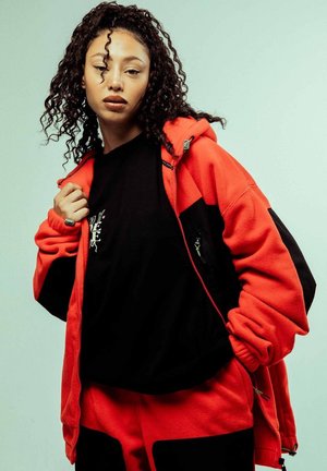 Chaqueta de forro polar roja y negra con un bolsillo con cremallera, usada sobre una camiseta negra. El modelo tiene el pelo rizado y posa frente a un fondo claro.