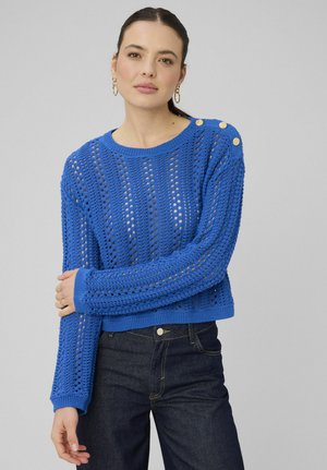 Femme portant un pull bleu en tricot avec des boutons dorés sur l'épaule et un jean foncé, debout devant un fond gris uni.