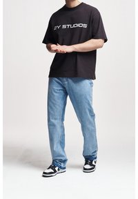 Camiseta negra de algodón con texto blanco "2Y STUDIOS", combinada con jeans de mezclilla azul claro y zapatillas negras/blancas. Corte casual.