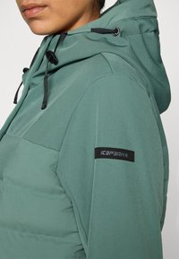 Zeleni puffa jakna s teksturiranim završetkom, koja ima kapuljaču, crne uže i logotip na rukavu.