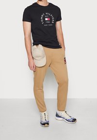 Černé bavlněné tričko s bílým logem, béžové tepláky s červenou výšivkou "H", šedá kšiltovka a vícebarevné sportovní boty.