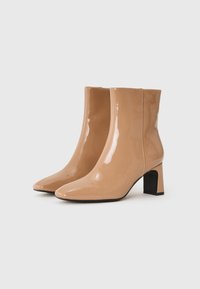 Botines de charol beige con punta afilada y tacón cuadrado. Acabado liso con costuras visibles a lo largo de las uniones.