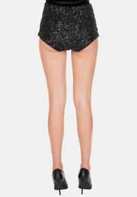 Donna con pantaloncini neri con paillettes a vita alta e scarpe nere con tacco alto, in piedi di spalle su uno sfondo semplice.