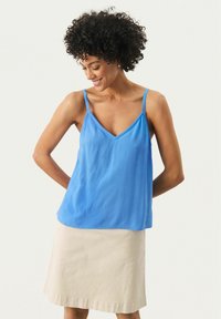 Camisola albastră din satin cu decolteu în V, bretele subțiri și o croială lejeră, purtată deasupra unei fuste beige deschisă până la genunchi. Textură netedă, ușoară.