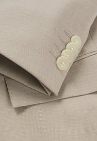 Tessuto beige strutturato con un motivo a scacchi, caratterizzato da quattro bottoni chiari e cuciture dettagliate sul polsino.