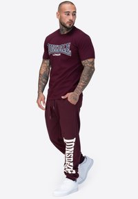 Burgunderfarbenes T-Shirt mit weißem Logo, kombiniert mit bordeauxfarbigen Jogginghosen mit weißem Druck. Getragen mit weißen Sportschuhen.