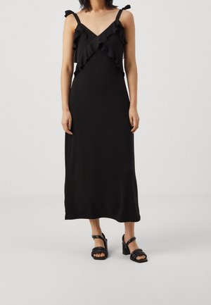 Femme portant une robe midi noire sans manches avec des détails volantés et des sandales à talons noires, debout devant un fond clair uni.