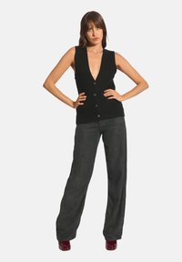 Černá bezrukávová vesta s hlubokým výstřihem do V, zapínáním na knoflíky a ribkovanou strukturou, zkombinovaná s volnými šedými širokými kalhotami a burgundskými botami.