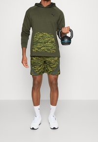 Olivenfarbener Hoodie mit Tarnmuster-Tasche, kombiniert mit passenden Shorts. Weiße Sneaker und eine schwarze Kettlebell mit blauen Akzenten.