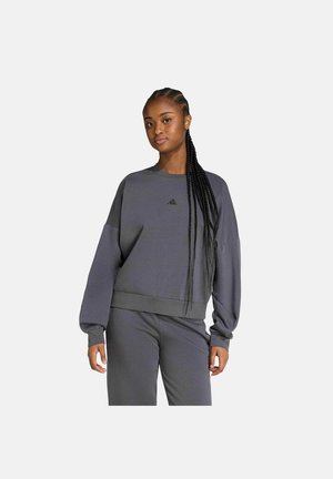 Frau mit langen Zöpfen, die einen lockeren grauen Adidas-Sweatshirt und passende Hosen trägt, vor einem schlichten hellen Hintergrund stehend.