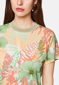 Blumenmuster T-Shirt in hellem Beige mit grünen und roten botanischen Designs, ausgestattet mit einem Rundhalsausschnitt mit kontrastierendem Saum und weichem Stoff.