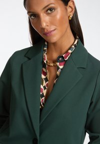 Groene getailleerde blazer van textuurstof, met sterke revers en een knoopsluiting, gecombineerd met een gebloemd blouse en een gouden ketting.