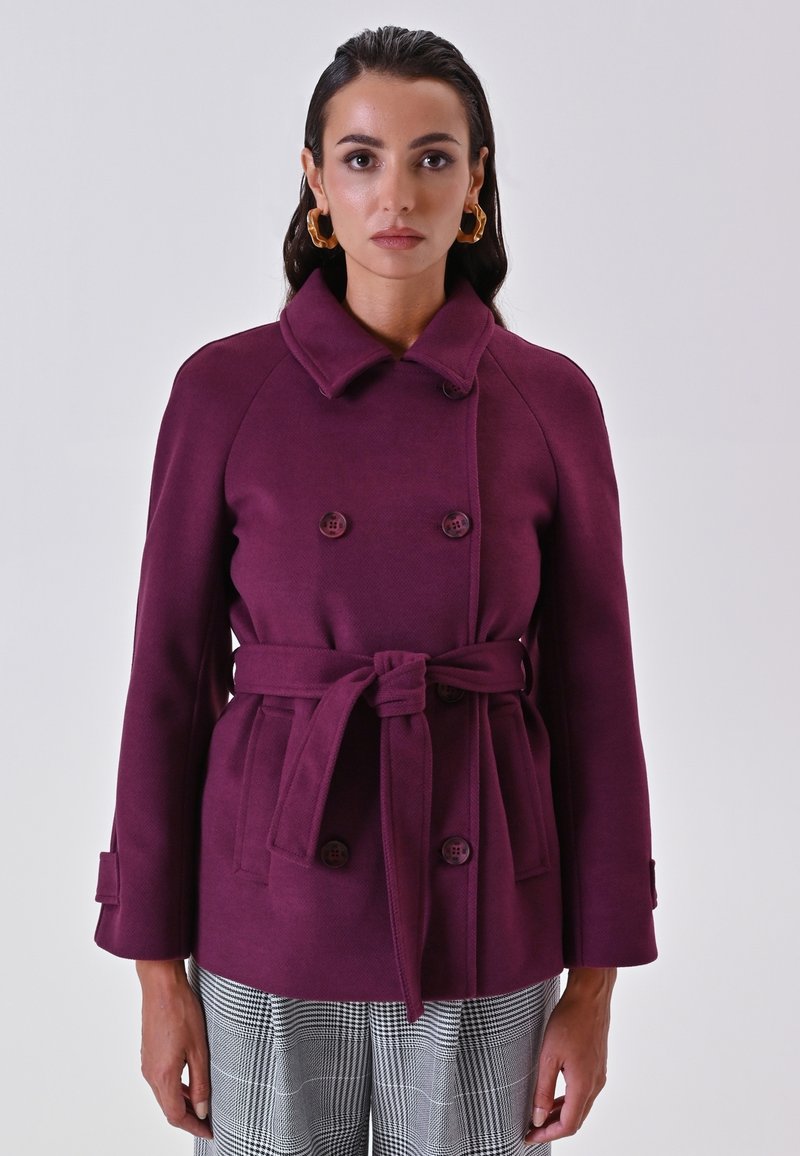 Cappotto bordeaux doppiopetto con cintura, tessuto texture, colletto a scollo a V e bottoni oversize. Indossato sopra pantaloni a quadri.