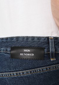 Vaqueros de mezclilla azul oscuro con un parche de cuero negro con el texto "WON HUNDRED." Costuras visibles y diseño clásico de cinco bolsillos.