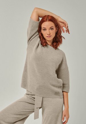 Junge Frau mit roten Haaren trägt einen beigen Ripp-Pullover und passende Hose, posiert mit einem erhobenen Arm vor einfarbigem Hintergrund.