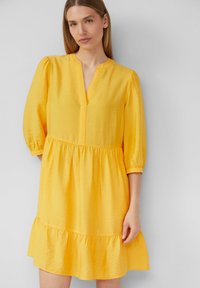 Gelbes Kleid mit V-Ausschnitt, Dreiviertelärmeln und einem gestuften Rüschensaum. Der Stoff hat eine strukturierte Oberfläche.