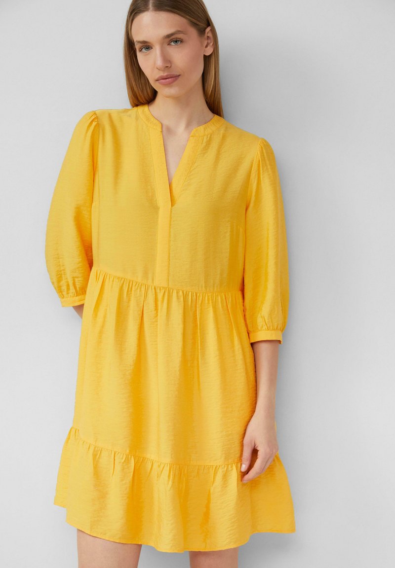 Gelbes Kleid mit V-Ausschnitt, Dreiviertelärmeln und einem gestuften Rüschensaum. Der Stoff hat eine strukturierte Oberfläche.