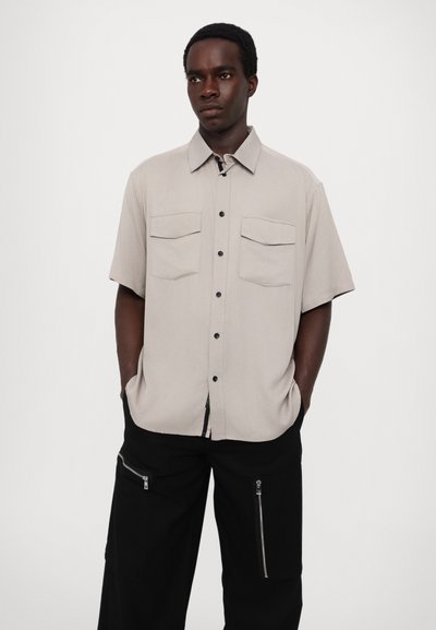 Chemise à manches courtes, de couleur gris clair, avec un devant à boutons, deux poches poitrine et un tissu respirant. Assortie à un pantalon cargo noir à zip.