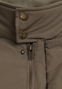 Chaqueta marrón acolchada con un alto cuello, botones de presión metálicos y una cremallera frontal con grabado de la marca. La tela parece ligera.