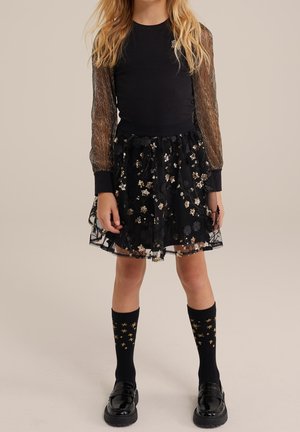 Fille portant une robe noire avec des manches transparentes ornées de fils métalliques et une jupe à sequins floraux, assortie de chaussettes noires et de mocassins brillants.