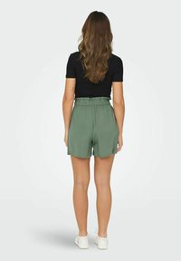 Calções verdes de cintura alta com um tecido texturizado e faixa elástica na cintura, combinados com uma blusa justa preta e sapatos brancos, vistos pela parte de trás.