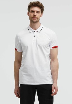 Man wearing a white short-sleeve polo shirt with black and red trim on sleeves and black pants against a plain background.