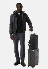 Donkergrijze bomberjack over een zwarte trui, grijze broek en zwarte schoenen. Grijze harde koffer met een zwarte leren rugzak erop.