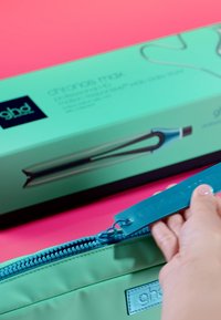 Hand, die einen türkisfarbenen Beutel neben einer passend dazugehörigen ghd-Haarglätter-Box auf einer rosa Oberfläche aufzippt.