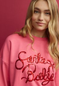 Rosa sweatshirt med en "Santa Baby" design i röda paljetter, med en avslappnad passform och mjuk textur. Bakgrunden är enfärgad rosa.