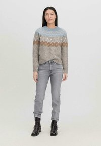 Neulepusu vaaleanharmaana, jossa on sininen ja ruskea kuviollinen kaulus. Yhdistetty vaaleanharmaisiin farkkuihin ja mustiin nilkkureihin, seisoo yksinkertaista taustaa vasten.