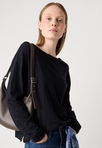 Woman with straight blonde hair wearing black long-sleeve shirt, blue jeans with a blue bandana, carrying a dark brown shoulder bag against a white background.