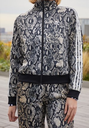 Person iført sort og beige Adidas træningsdragt med slangeprint og hvide tre-stribe-detaljer på ærmer og bukser udendørs.