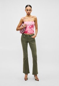 Γυναίκα με ροζ φλοράλ τοπ χωρίς τιράντες, ελαιοπράσινο τζιν με φαρδιά μπατζάκια, πράσινα πέδιλα με ψηλό τακούνι και κρατώντας ροζ τσάντα με παγιέτες.