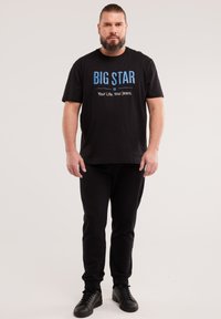 Uomo con barba e capelli raccolti, che indossa una maglietta nera con la scritta "BIG STAR", pantaloni neri e scarpe da ginnastica nere, in piedi su sfondo semplice.
