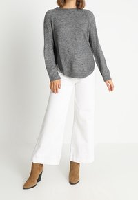 Pullover grigio a maniche lunghe con orlo arrotondato, abbinato a pantaloni bianchi a gamba larga e stivali con tacco medio color cammello. L'outfit presenta un maglione lavorato a maglia testurizzata.