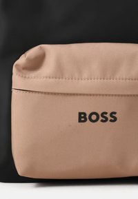 Bežinė audinio krepšio kišenė su tekstūruotu paviršiumi, turinti juodą "BOSS" logotipą ir užtrauktuką, esančią juodoje fone.
