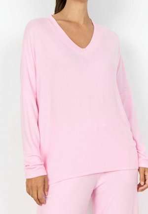Femme portant un haut rose pâle à manches longues avec col en V et un pantalon assorti, montrant le haut du corps et une partie des jambes sur fond blanc uni.