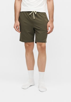 Férfi, aki oliva zöld, húzózsinóros rövidnadrágot, fehér zoknit és fehér felsőt visel, sima fehér háttér előtt áll.