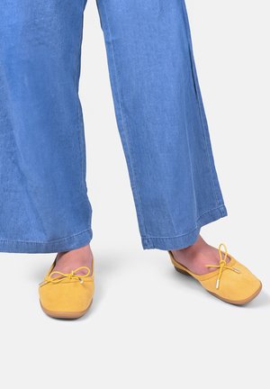 Stopy w żółtych, kwadratowych butach wsuwanych z kokardkami, w połączeniu z niebieskimi, szerokimi spodniami na tle jednolitego tła.