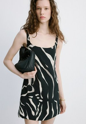 Jong persoon met lang krullend haar die een zwart-witte zebra gestreepte jurk draagt, een zwarte geweven handtas vasthoudt en een gouden armband om de pols draagt.