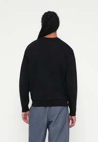 Sweatshirt preto com decote redondo, punhos e bainha canelados, apresentando uma textura suave. Usado com calças de ganga azuis.