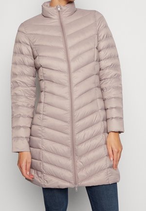 Vrouw die een licht mauve, doorgestikte lange donsjas draagt met een voorrits en ontspannen met haar handen langs haar zij staat.