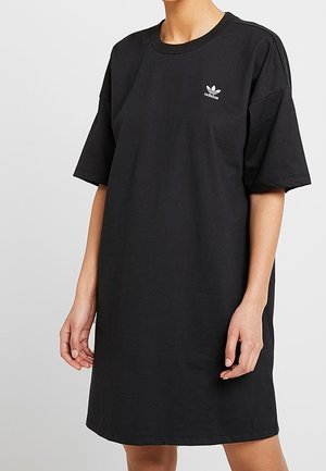 Vrouw draagt een losse zwarte Adidas T-shirtjurk met korte mouwen en een klein wit logo op de borst tegen een effen achtergrond.
