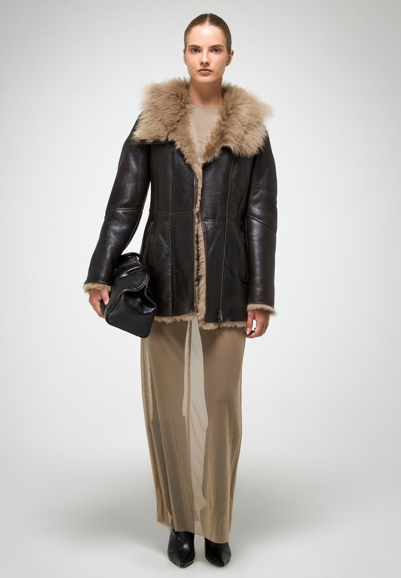 Tumši ādas jaka ar bēša krāsas kažokādas apkakli, ar rāvējslēdzēju priekšā, divām sānu kabatām, apvienota ar garu, caurspīdīgu bēša krāsas svārku un melnu apavu.