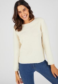 Pull en tricot couleur crème avec un col rond, des accents texturés sur le devant et des manches longues, associé à un jean en denim.