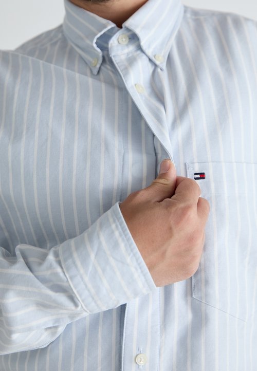 Chemise boutonnée à rayures bleu clair avec col à boutons, poche poitrine simple et accent de logo sur la poche. Texture de tissu doux.