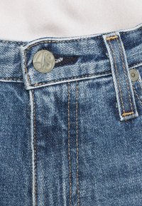 Džínové kalhoty s knoflíkovým zapínáním, obsahující stříbrný knoflík s rytinou „49“, modrou látku se zlatou nití a opotřebovaný vzhled.