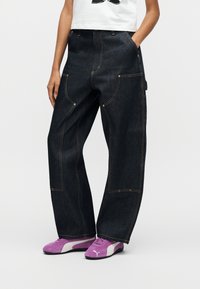 Άτομο που φοράει σκούρο μπλε φαρδιά τζιν παντελόνια με πορτοκαλί ραφές, λευκό κοντό τοπ και μοβ αθλητικά παπούτσια Puma, στέκεται με τα χέρια στις τσέπες.