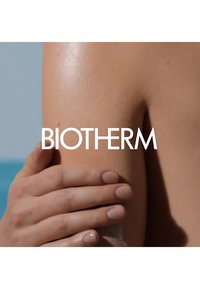 Gros plan sur un bras nu avec une peau lisse, une main appliquant une crème. L'arrière-plan est bleu, mettant en valeur l'éclat naturel de la peau. Le texte "BIOTHERM" est visible.